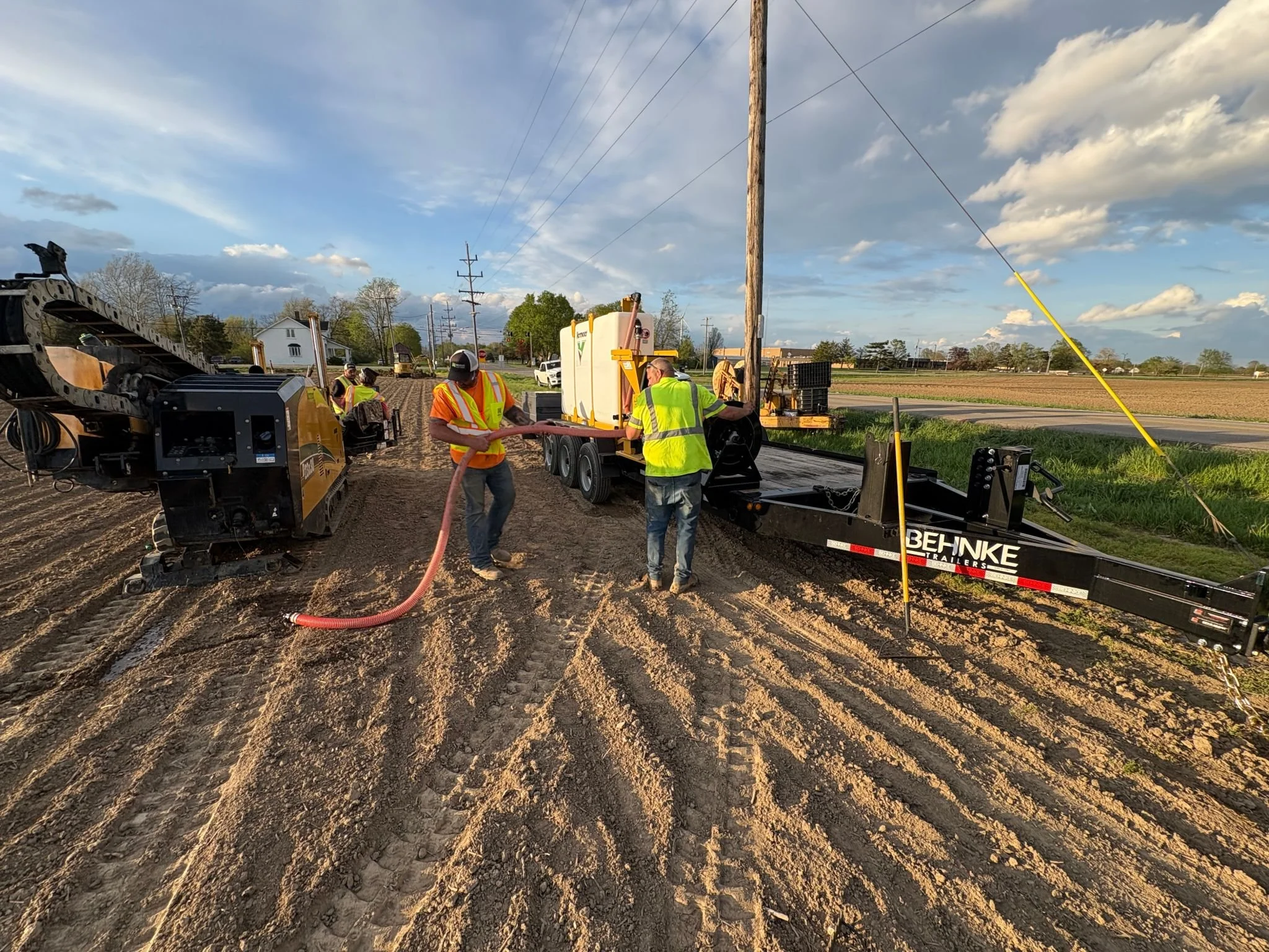 Construction workers in safety vests operate heavy equipment and machinery on a dirt road during daylight, with a background of a rural landscape and power lines.