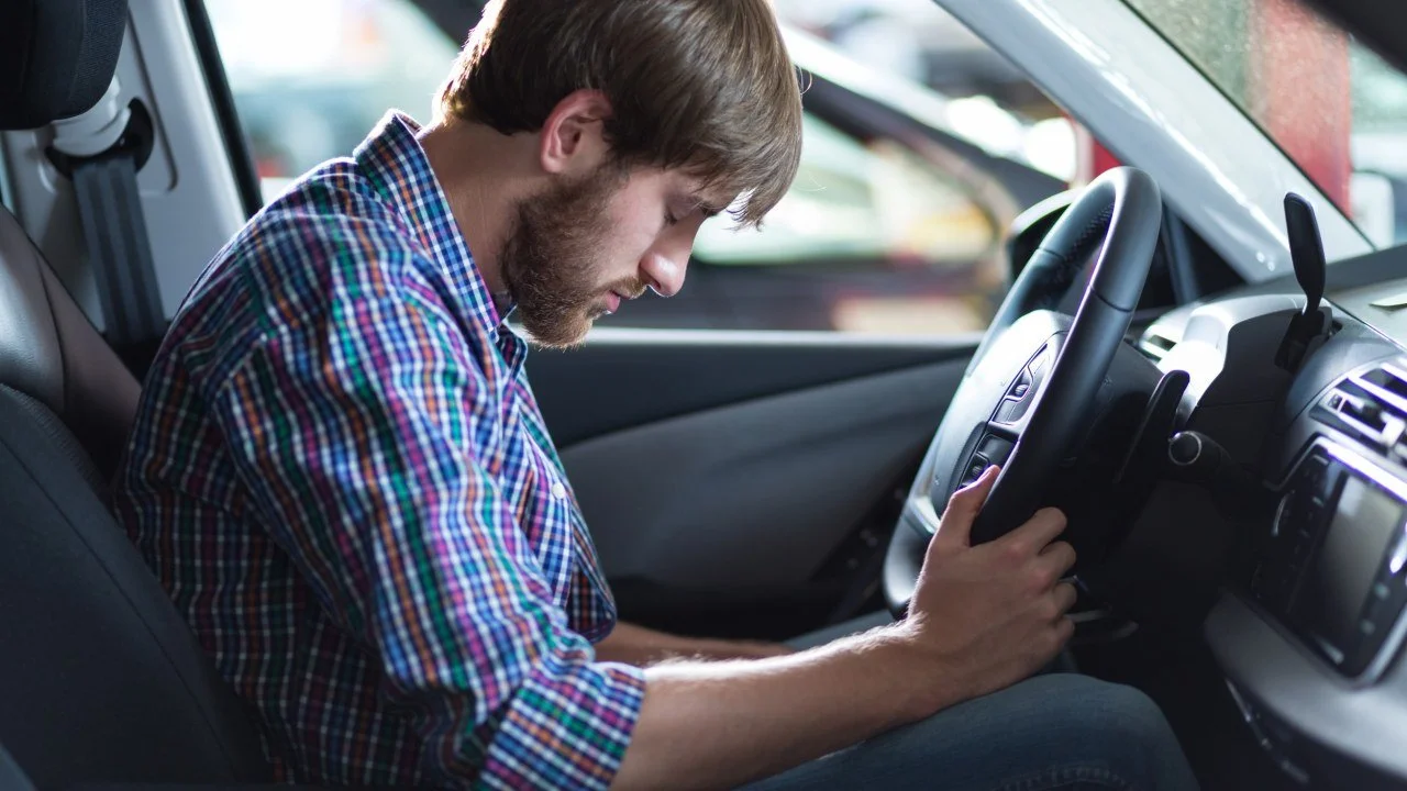 A young man with a beard and wearing a plaid shirt sitting inside a car, leaning his head on the steering wheel with eyes closed, appearing to be tired or sleeping.