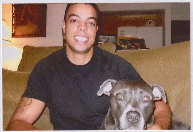A man smiling and sitting on a couch with a gray dog in a living room.