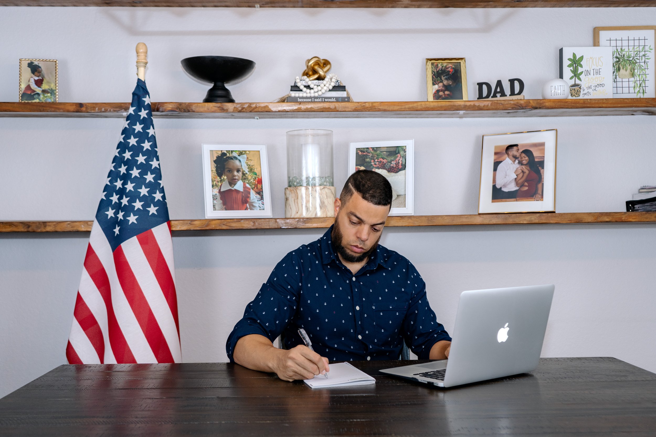 A man sitting at a dark wooden desk, writing in a notebook while working on a silver MacBook, with an American flag next to him, in a room decorated with family photos and small decor on wooden shelves.