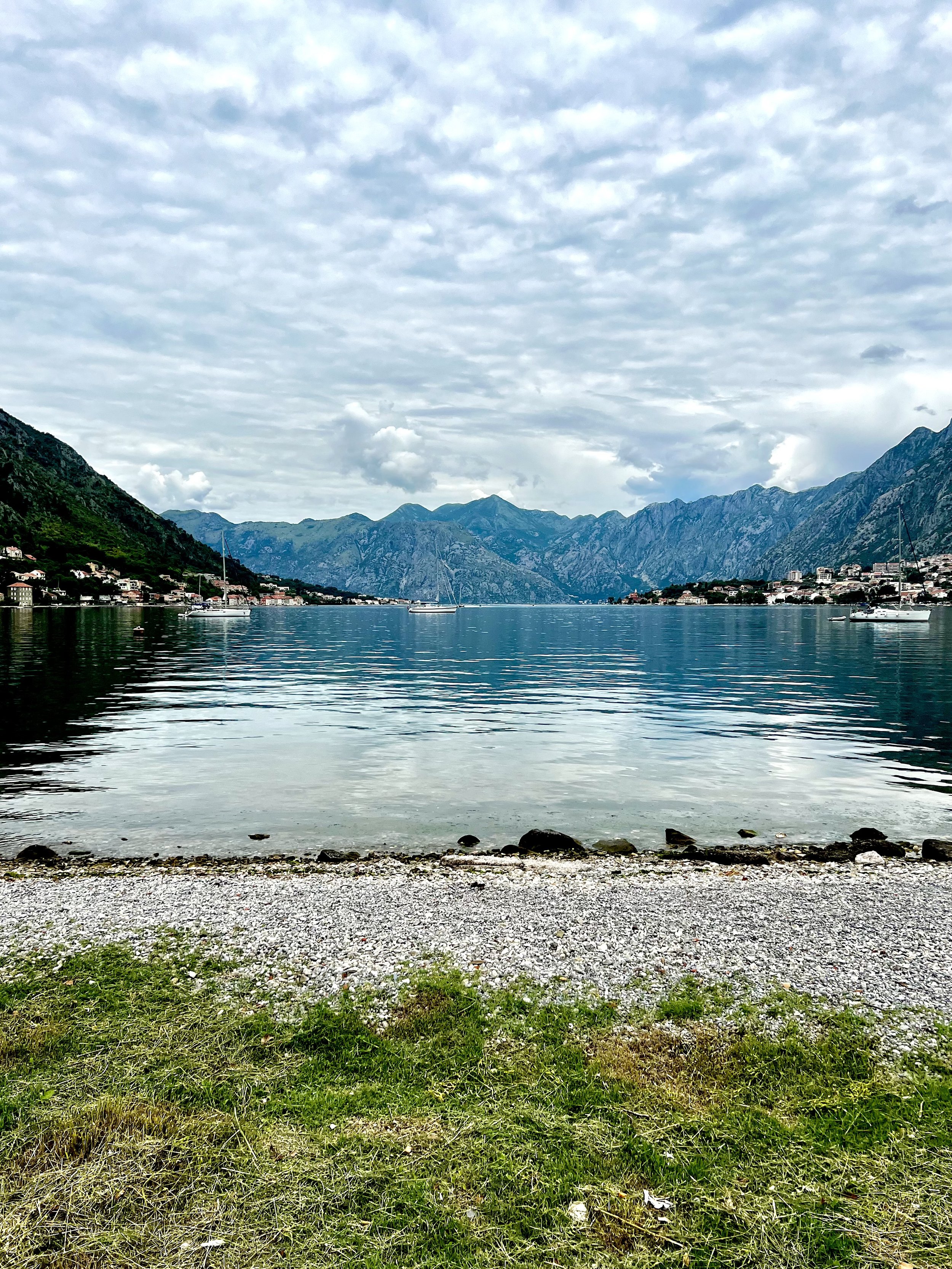 Scenic view of a calm bay with mountains in the background, cloudy sky, green grass, and rocky shoreline in Kotor Bay, Montenegro.