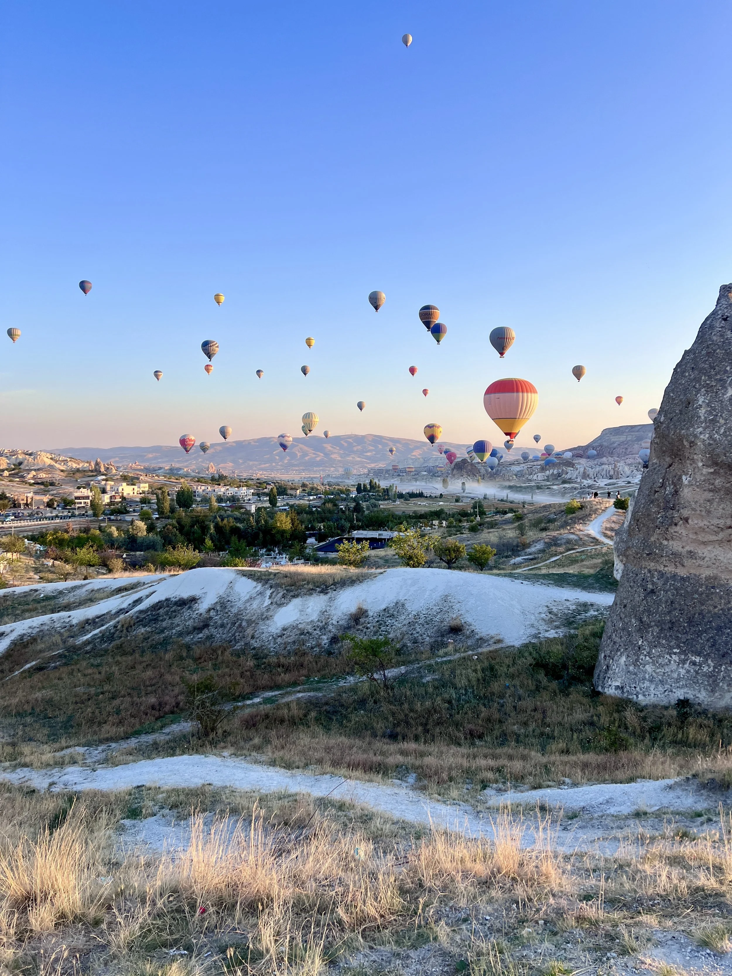 Hot air balloons floating over a scenic landscape with rolling hills and scattered vegetation in Cappadocia, Turkey.