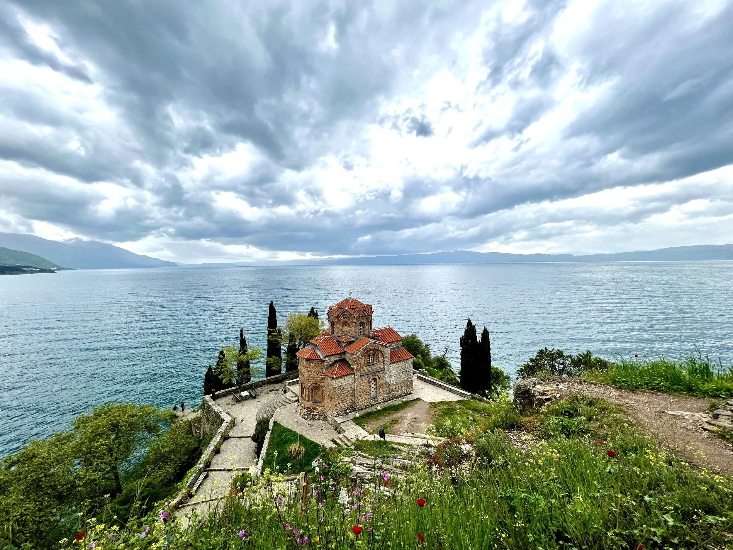 Scenic view of a historic church with red-tiled roof and stone walls, located on a hillside overlooking a large, calm lake. The sky is cloudy, and there are trees and colorful wildflowers surrounding the church in Ohrid, North Macedonia.