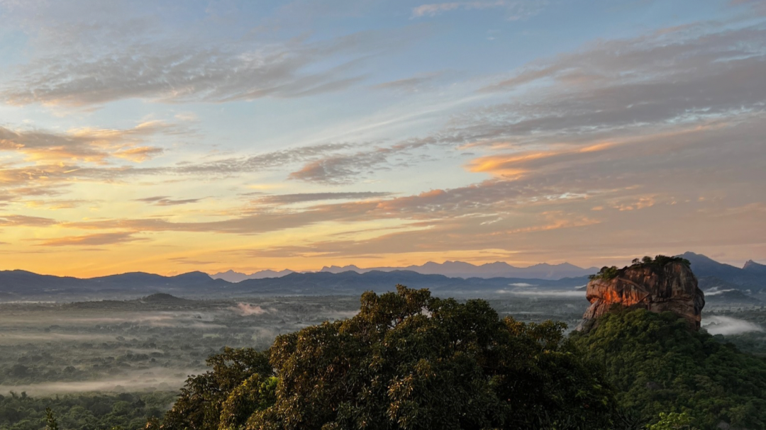 A colourful sunrise looking over Lions Rock in Sigirya, Sri Lanka with mountains and hills in the background and trees and the rock in the foreground