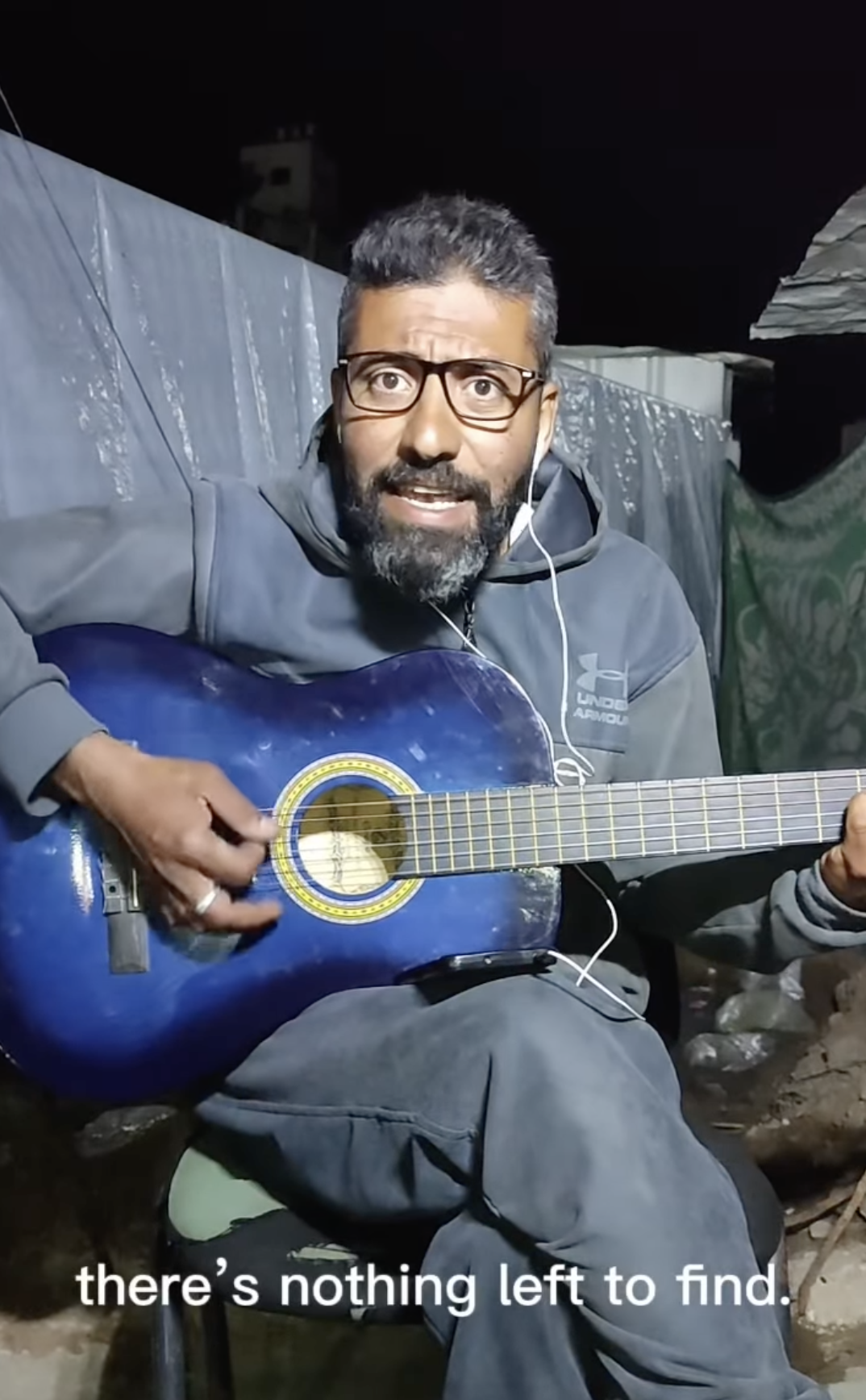 Musician in Gaza playing a blue acoustic guitar and singing, wearing glasses and headphones, with a tent behind him at night. Subtitles read "there’s nothing left to find."