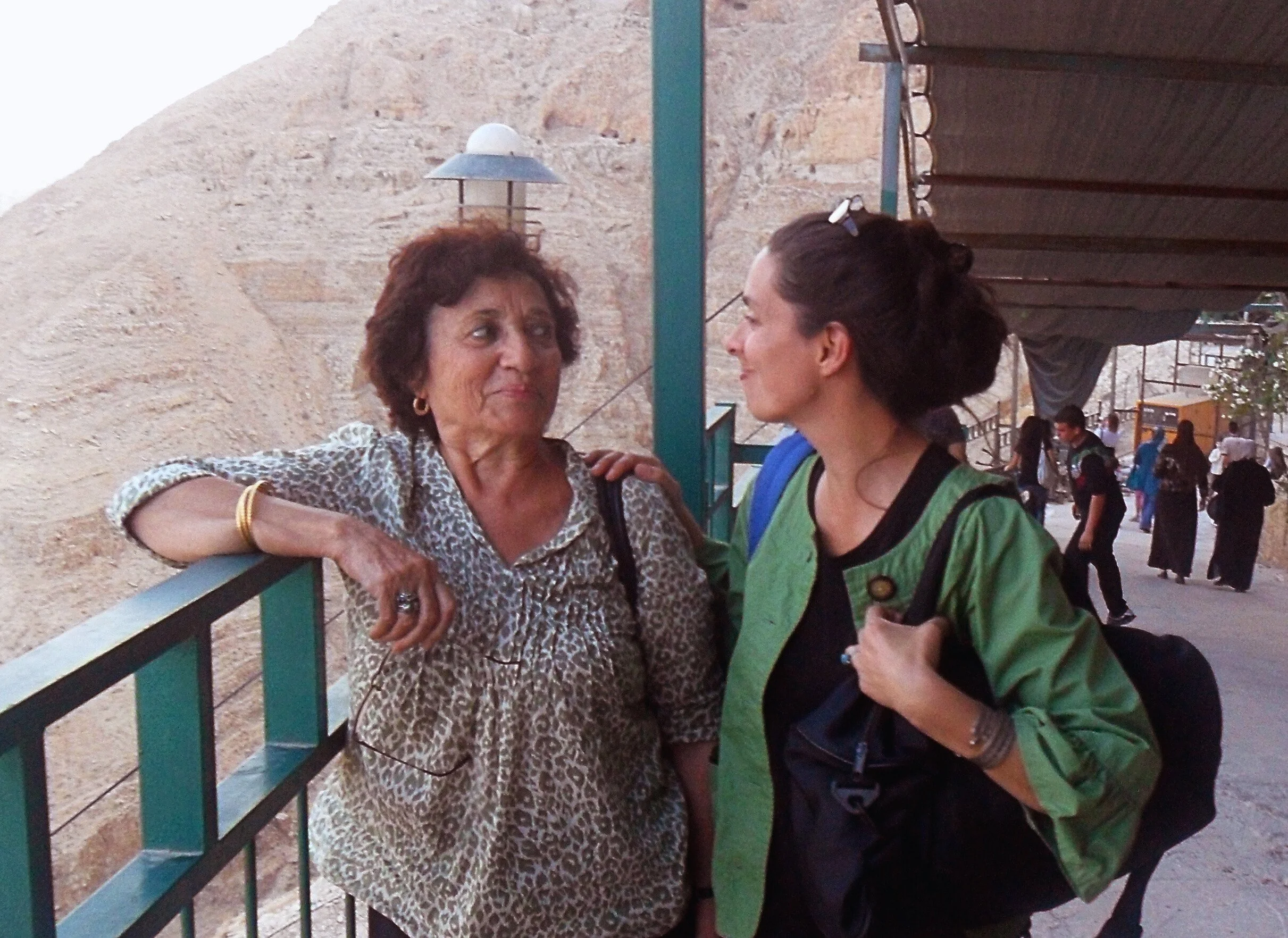 Two women standing on a terrace in a Jericho, West Bank, talking and smiling. One woman wears a leopard print blouse and the other a green jacket. People walking in the background.