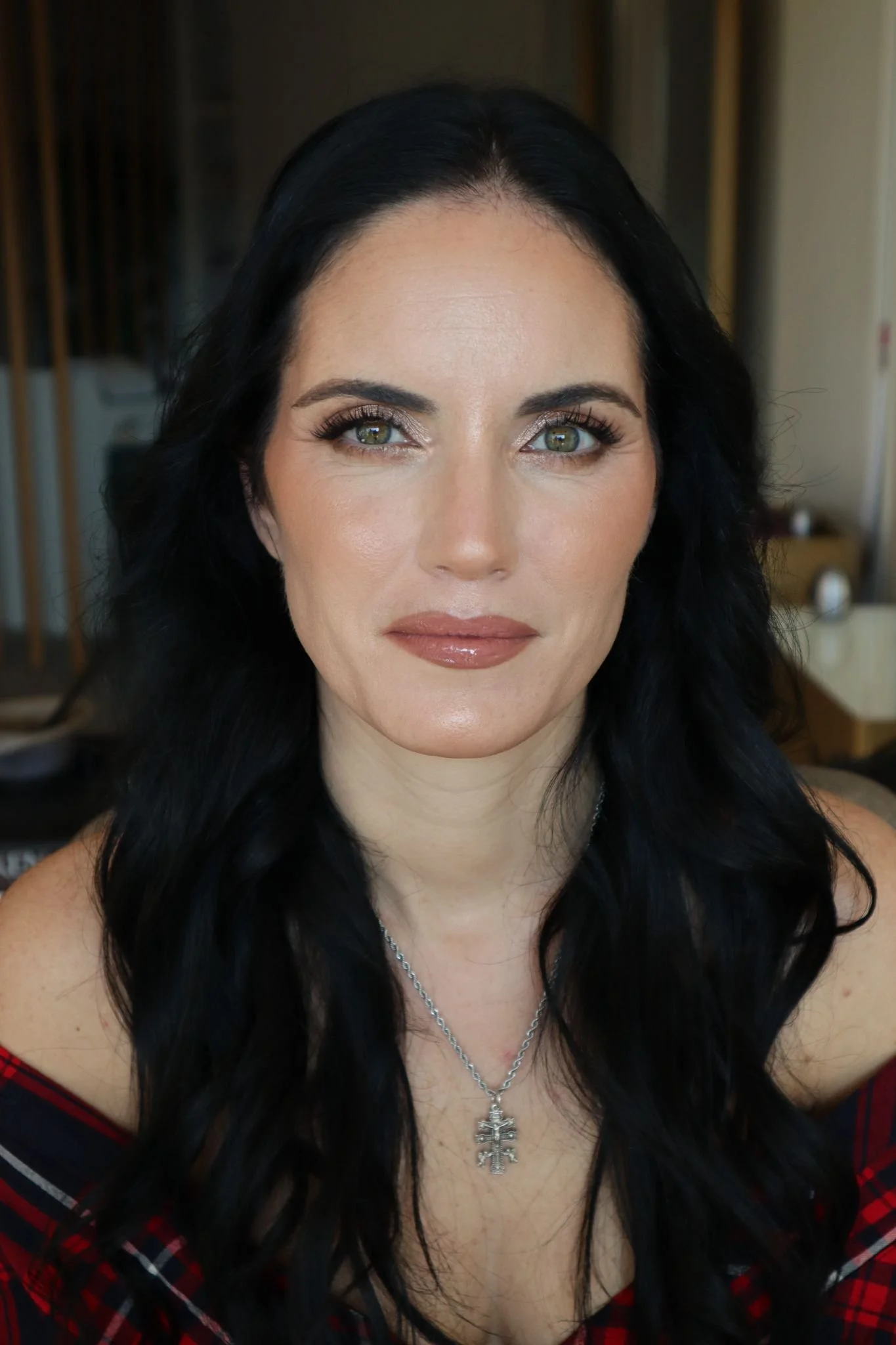 Mulher de cabelos pretos e longos, usando colar prateado, com expressão neutra, em ambiente interno.