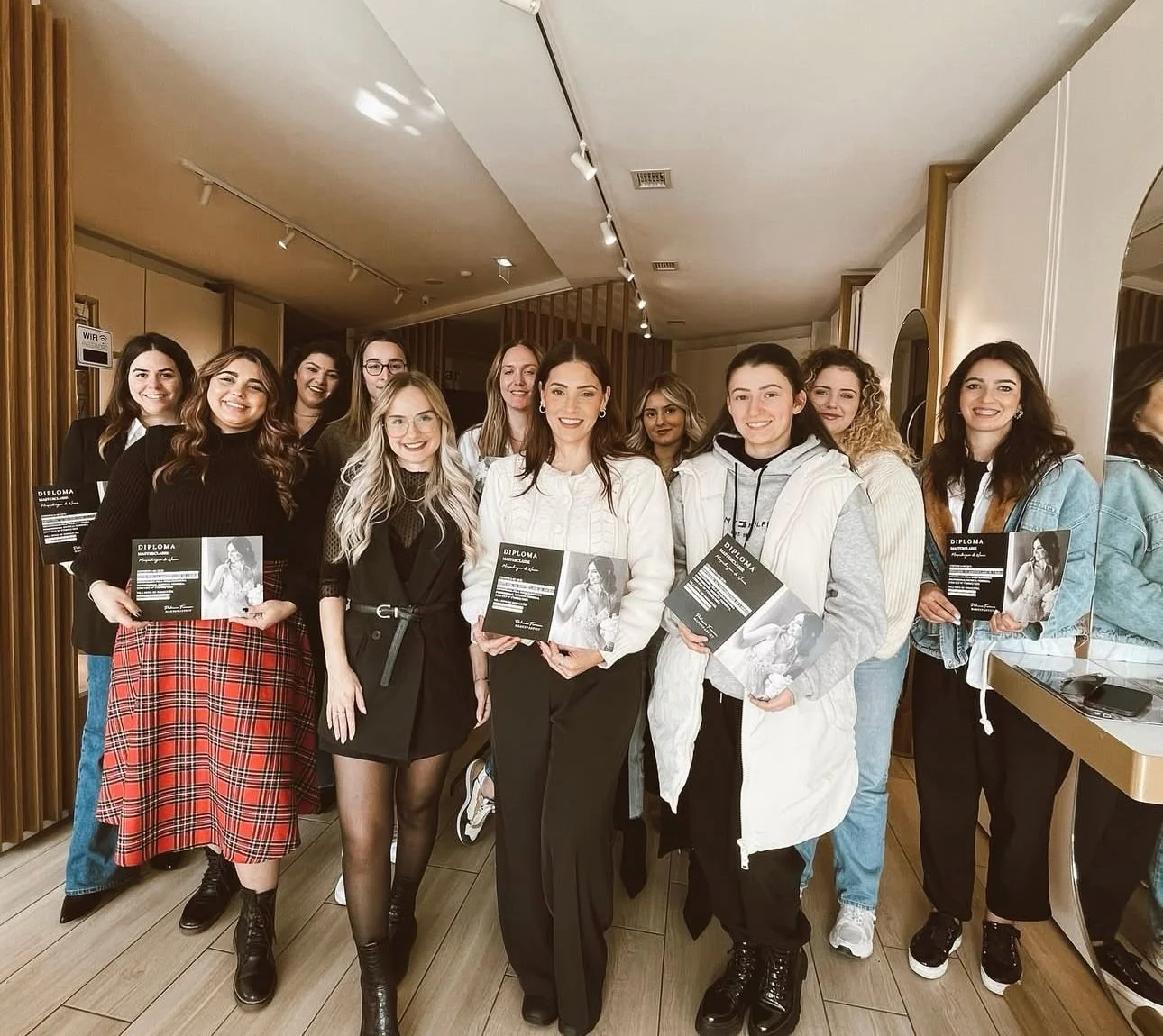 Grupo de mulheres posando para foto em ambiente interno, segurando diplomas. Elas estão sorrindo e vestem roupas diversas. A sala tem decoração moderna.