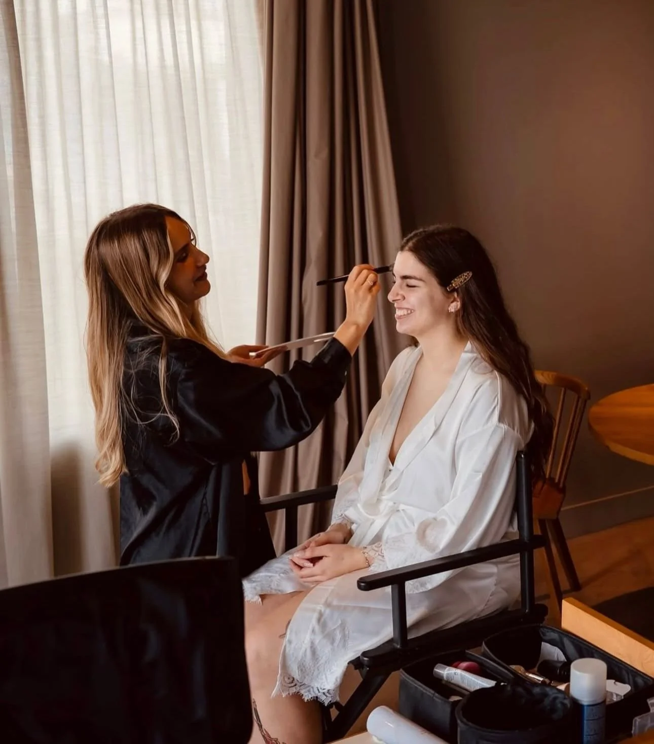 Uma mulher maquiando outra que está sentada em uma cadeira, usando um robe, em um ambiente interno com cortinas.