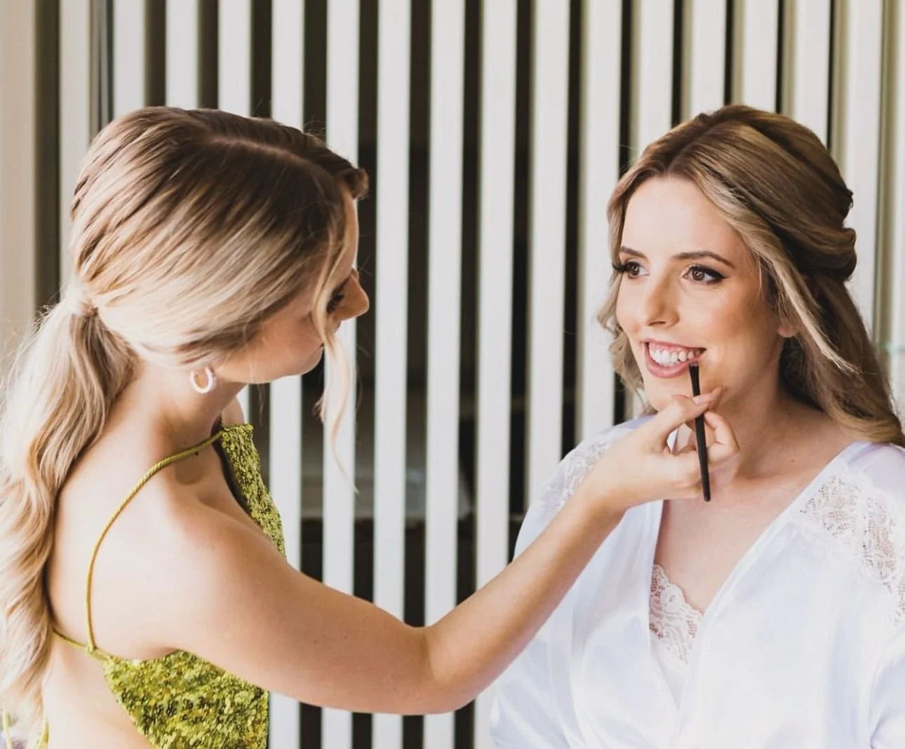 Duas mulheres, uma maquiando a outra, com um pincel nos lábios. A mulher sentada sorri, vestindo um robe branco. A outra usa um vestido verde brilhante. Fundo com persianas.