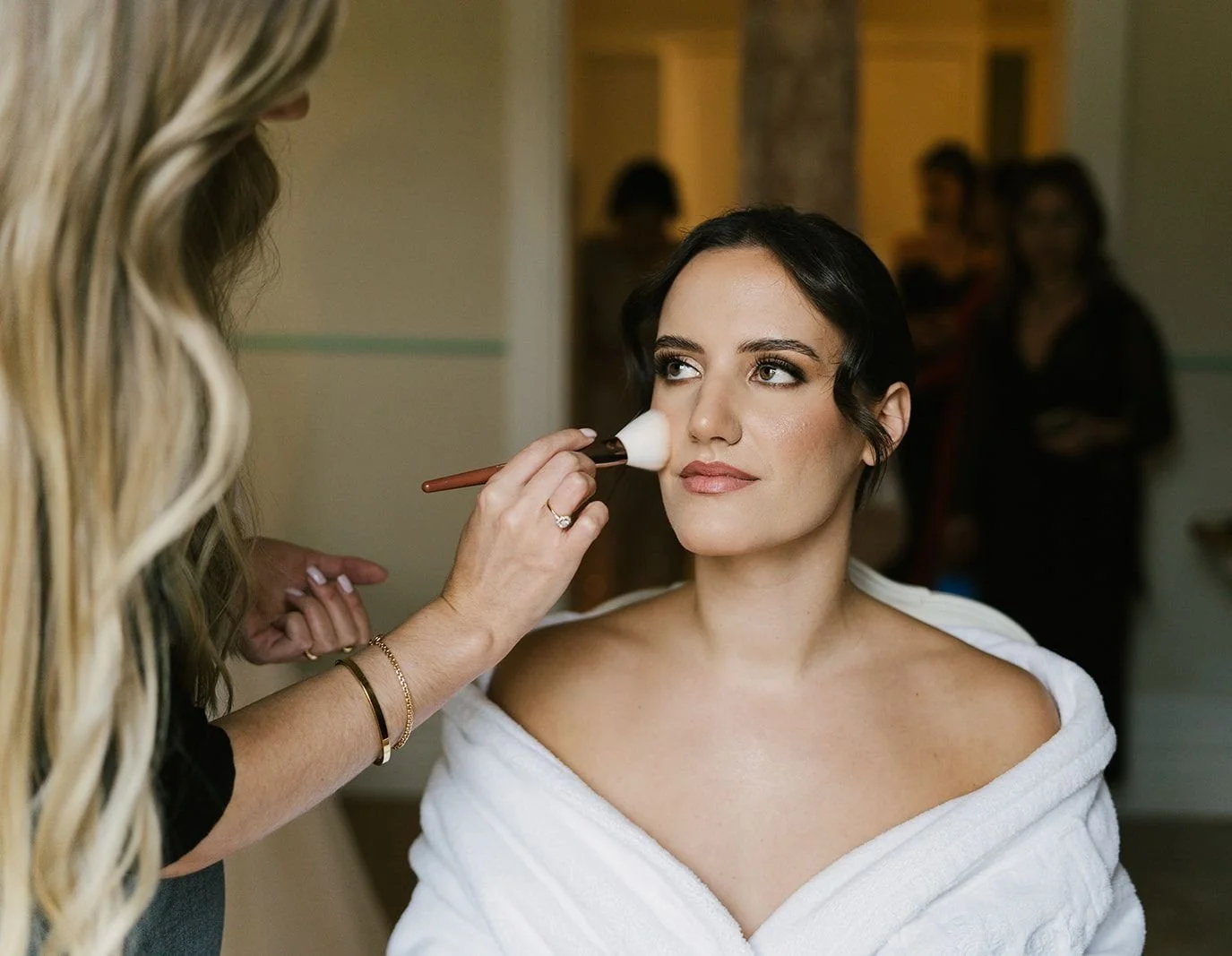 Uma noiva está a ser maquilhada. A Patricia Ferreira aplica o blush com um pincel. O ambiente é da preparação de um casamento.