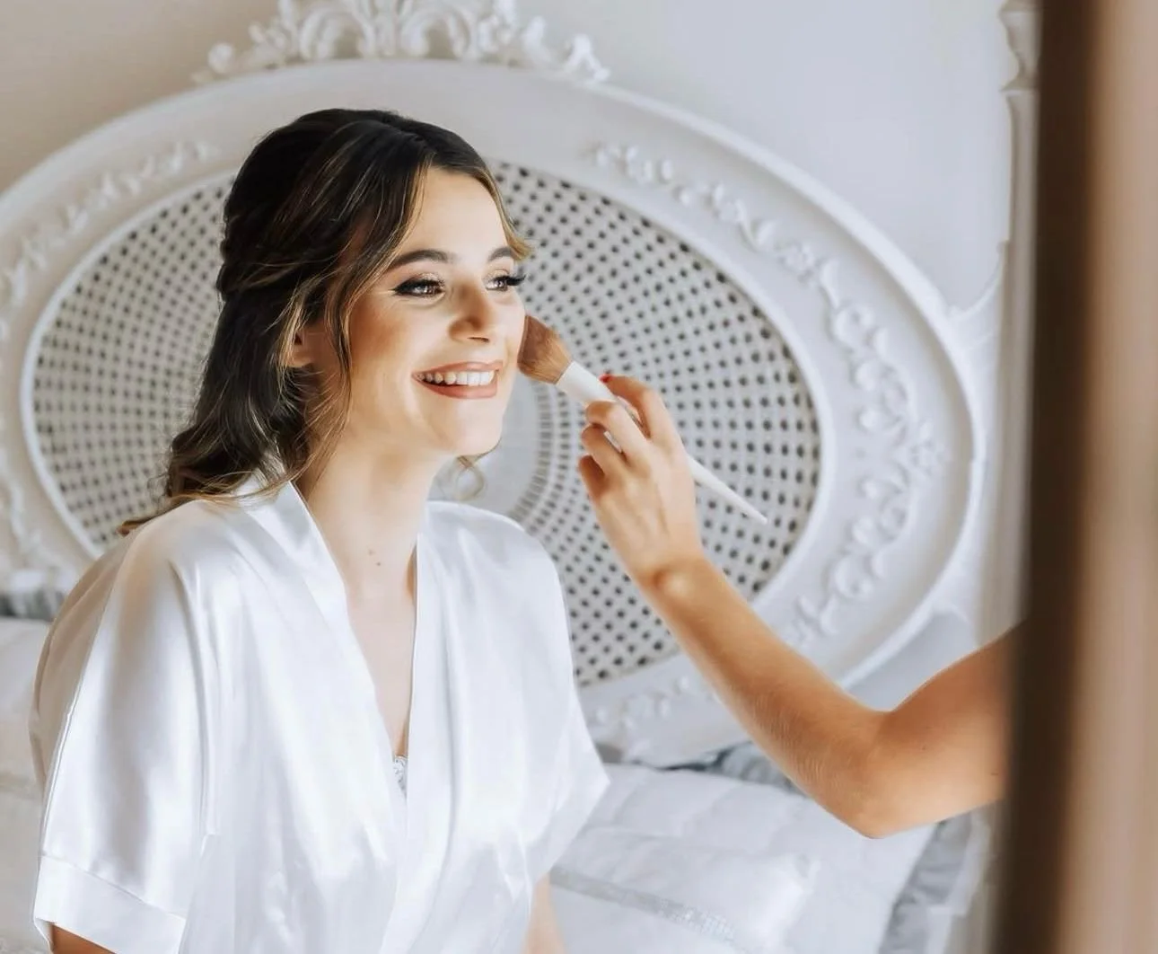 Mulher sorrindo sentada em uma cama enquanto alguém aplica maquiagem no seu rosto com um pincel.
