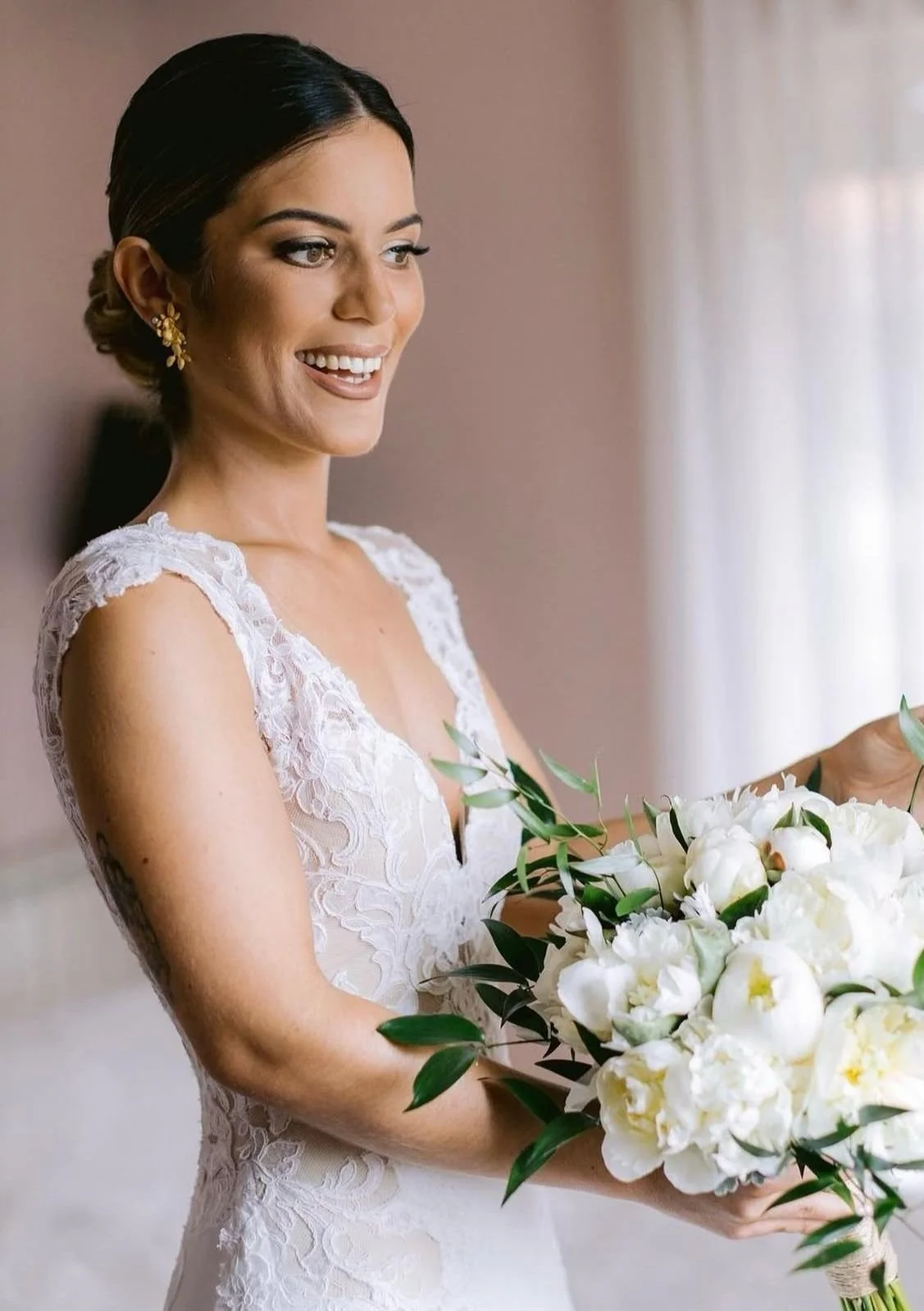 Noiva sorridente segurando um buquê de flores brancas, vestindo um vestido de renda branco.