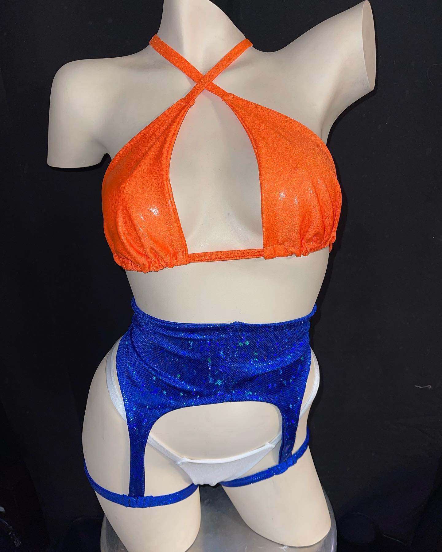 Mannequin wearing an orange bikini top with a halter neck and a blue sequined garter belt over white underwear, set against a dark background.