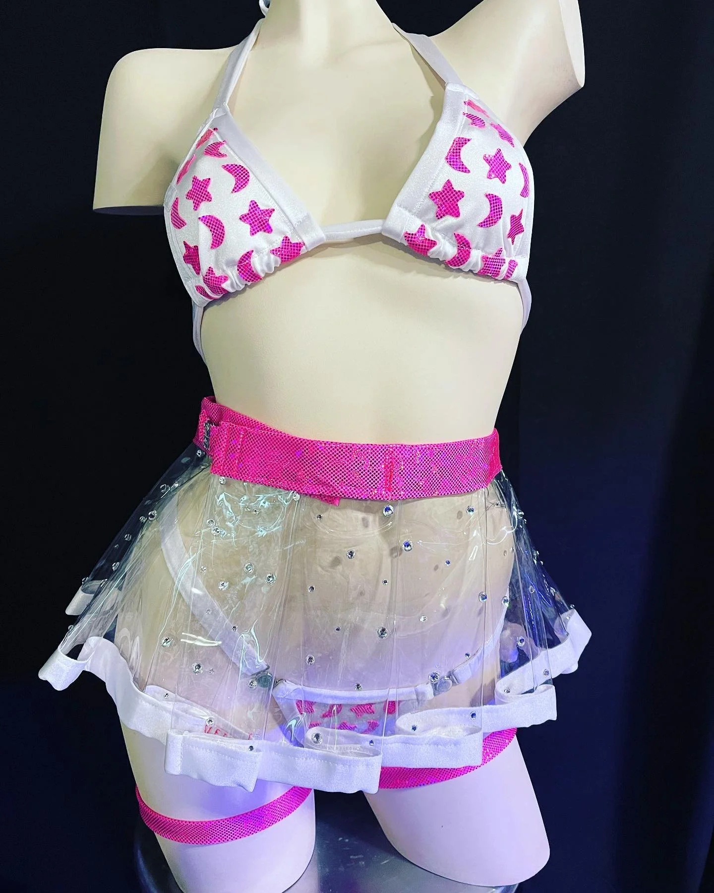 Mannequin wearing a pink and white bikini top with star and moon patterns, and a transparent skirt with sequins and pink waistband, set against a black background.