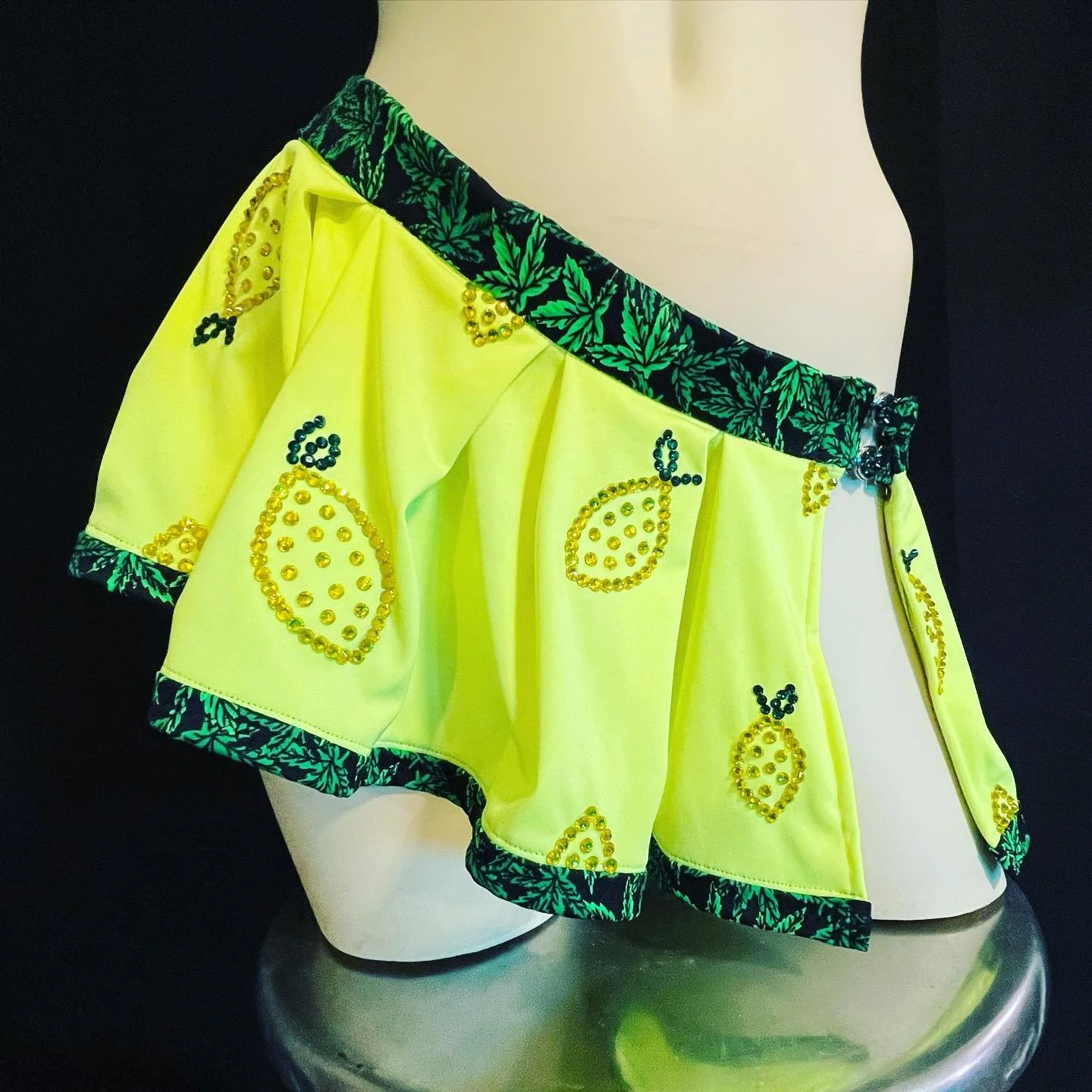 A yellow fabric with pineapple patterns and rhinestone embellishments draped over a mannequin or stand, featuring a decorative green leafy trim.