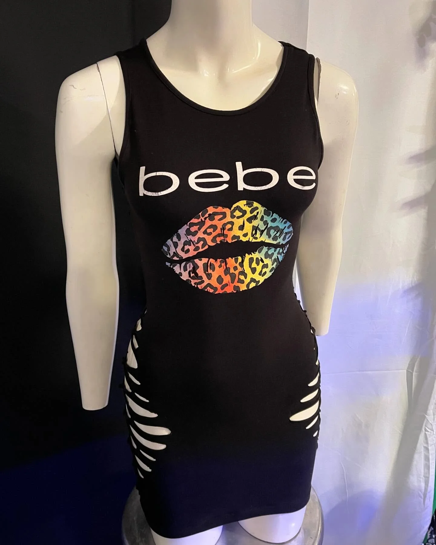 Mannequin wearing a black sleeveless dress with 'bebe' logo and colorful leopard print lips design. The dress features side cutouts.