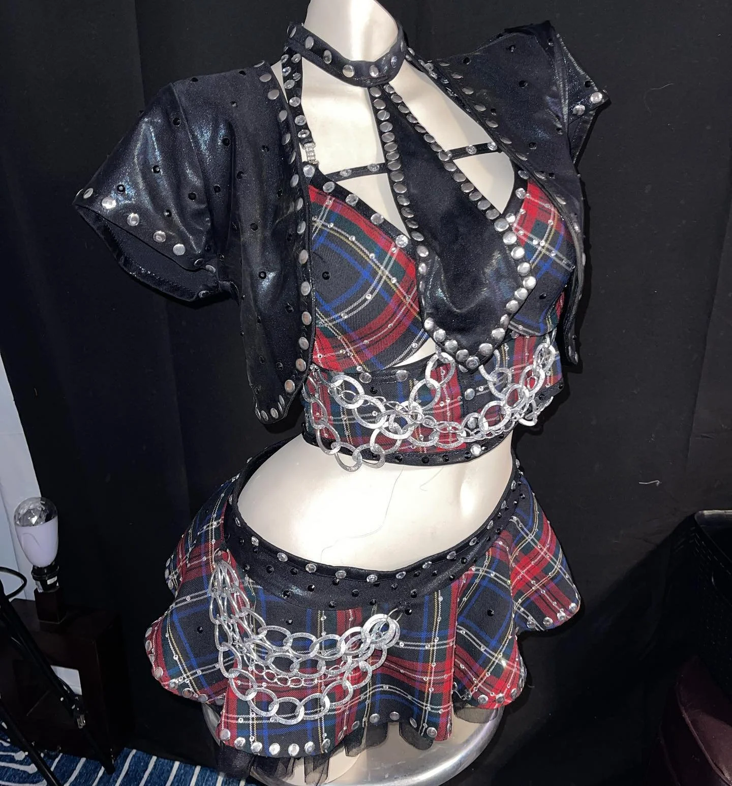 Mannequin wearing a black and red plaid outfit with metal chains and studded accents in front of a dark background.