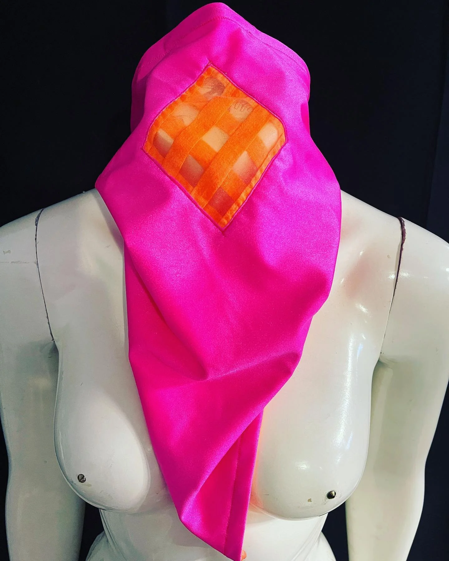 Mannequin wearing a bright pink headscarf with orange checkered detail, against a black background.
