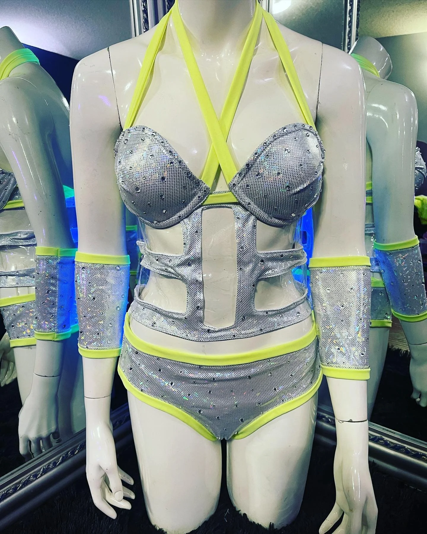 Mannequin wearing a silver studded outfit with neon yellow trim, featuring a halter top with cut-outs, matching bottoms, and arm sleeves, displayed beside a mirror.