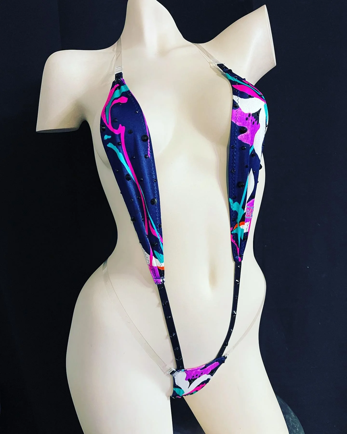 A mannequin wearing a colorful, abstract-patterned one-piece swimsuit with clear straps, showcased against a dark background.