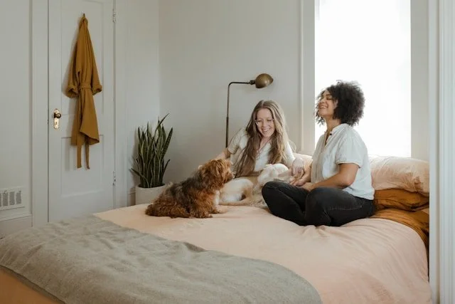 Two women sitting on a bed with two dogs, smiling and playing together in a bright bedroom.