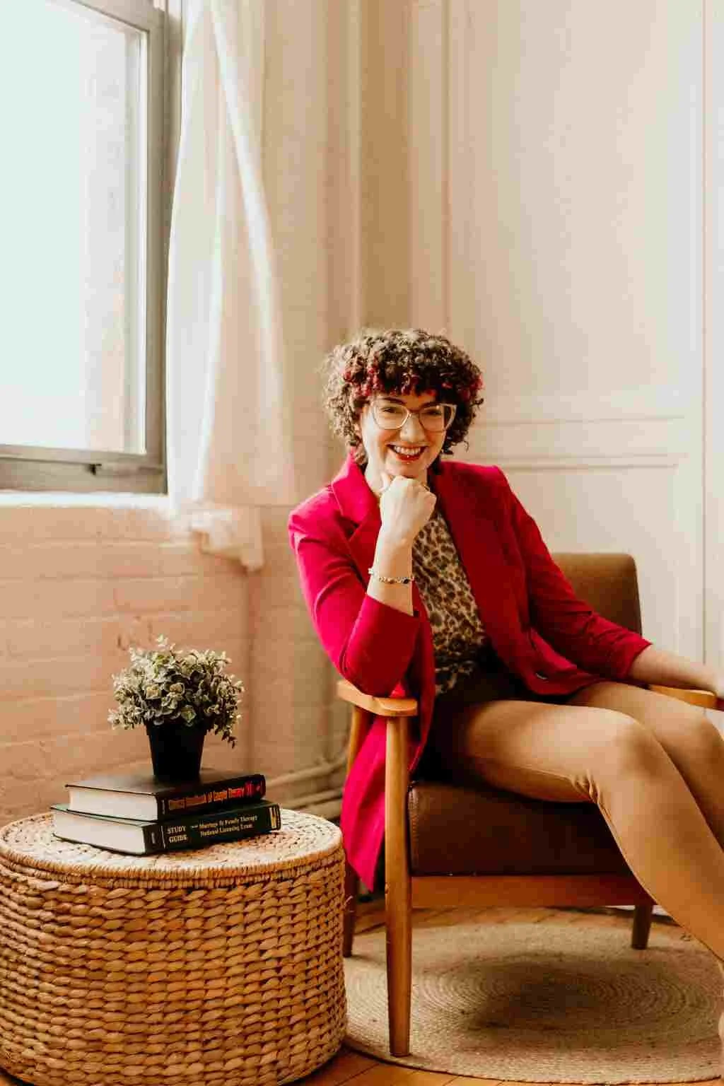 A woman with curly hair, glasses, and a bright smile, sitting comfortably in a modern room. She is wearing a blazer and tan pants, with a leopard-print top. Beside her is a woven round side table with a small black pot of flowers and several stacked books. The room has natural light coming through a window with white curtains.