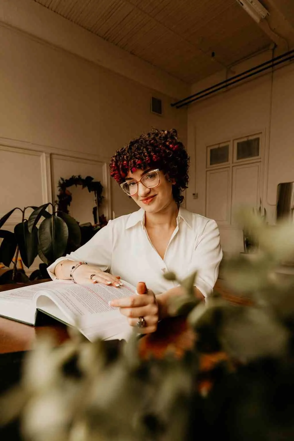 A woman with curly hair and glasses, sitting at a table with an open book, in a cozy room decorated with plants and a wreath on the wall.