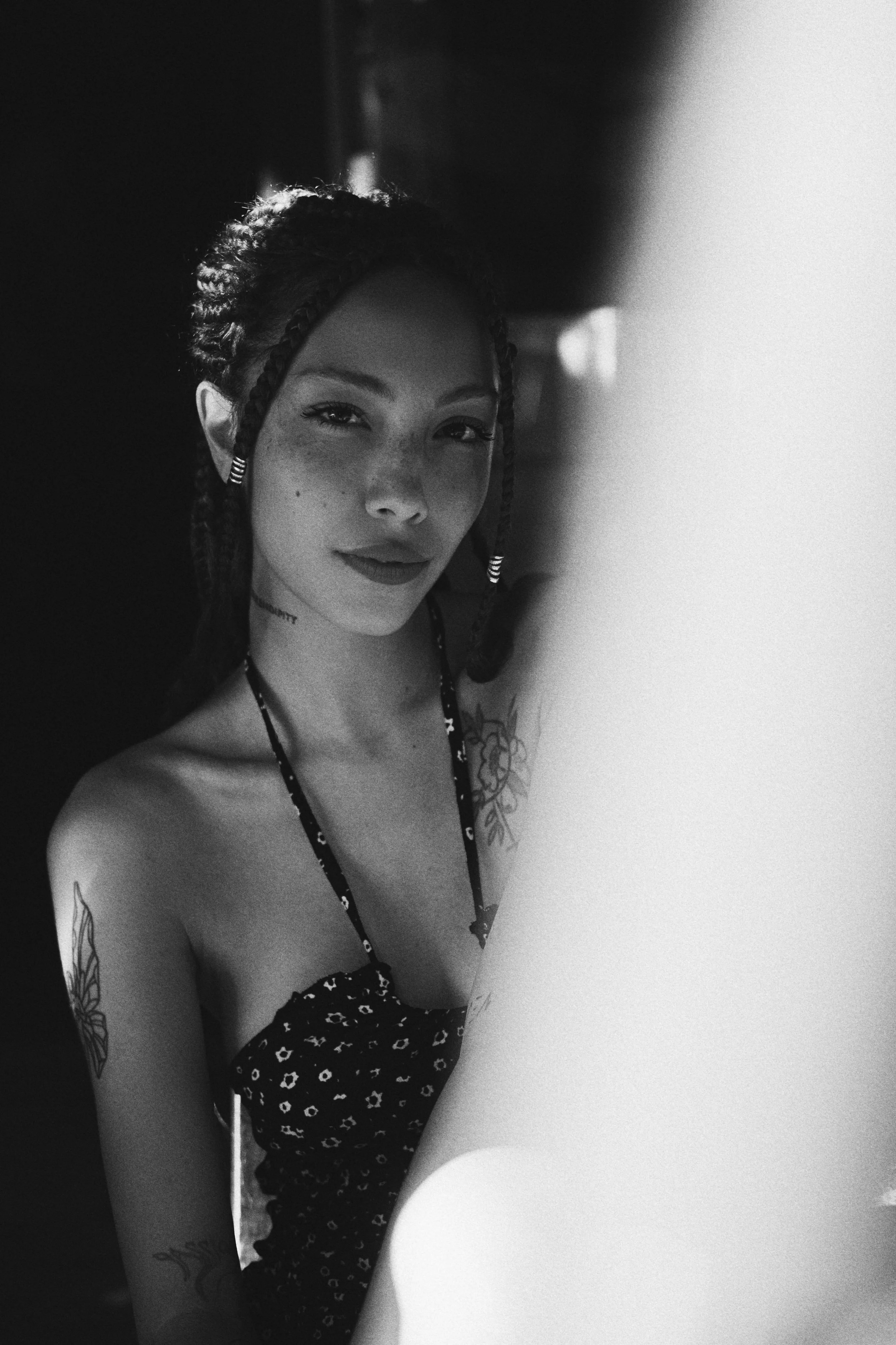 Black and white portrait of a person with braided hair, wearing a patterned dress, and showing tattooed arms.