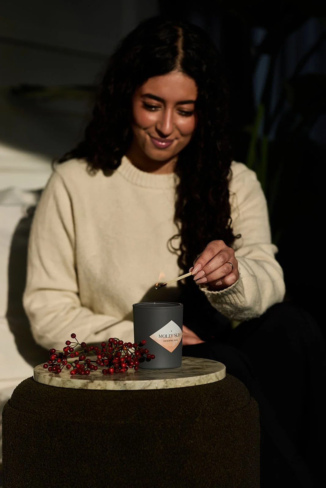 Eine junge Frau mit lockigem dunklem Haar, die eine Streichholzschachtel anzündet, sitzend neben einer Kerze und roten Beeren auf einem Tisch