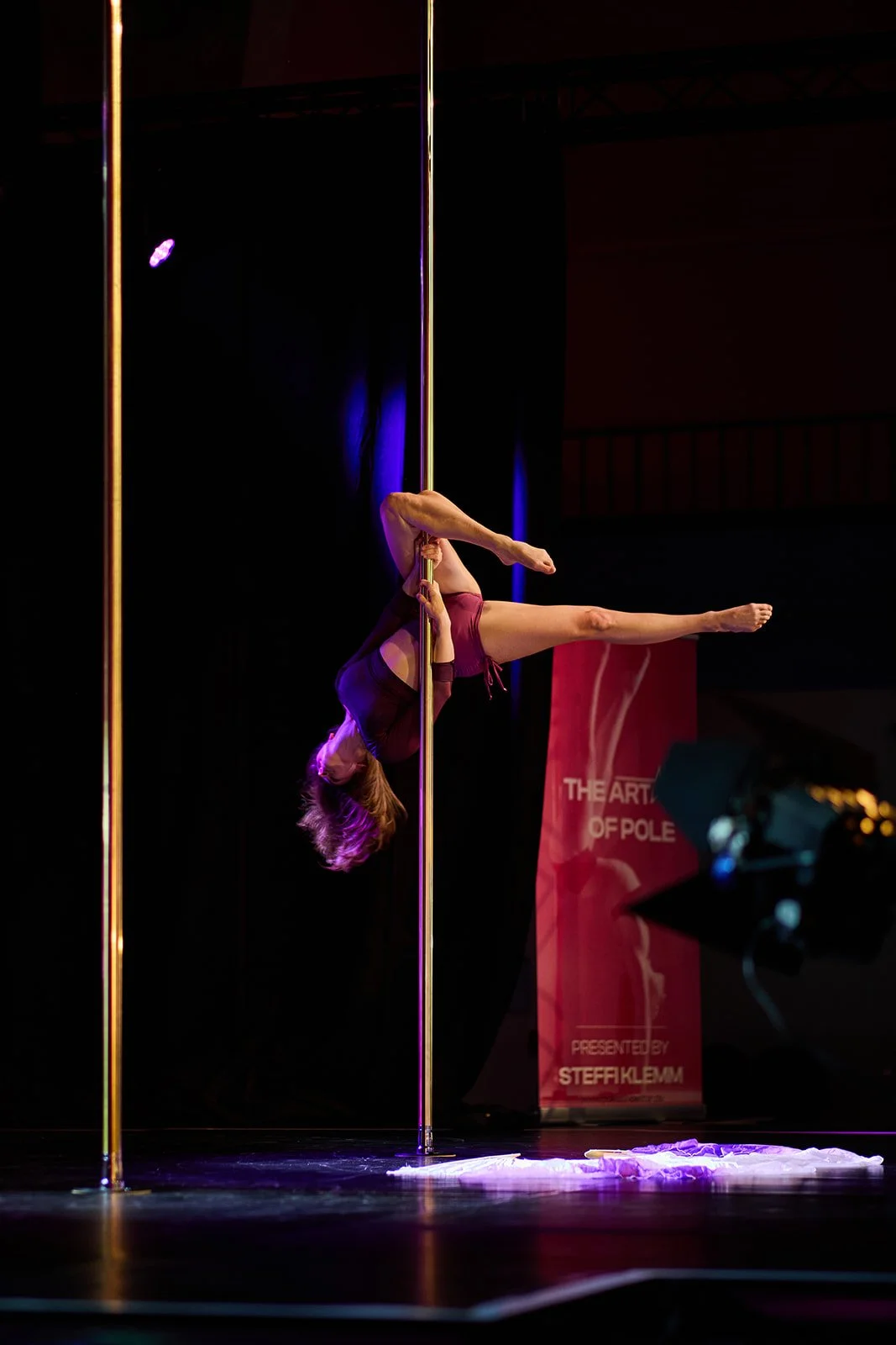 Eine junge Frau tanzt an einer vertikalen Stange auf der Bühne, mit dunklem Hintergrund und einem roten Banner im Hintergrund.