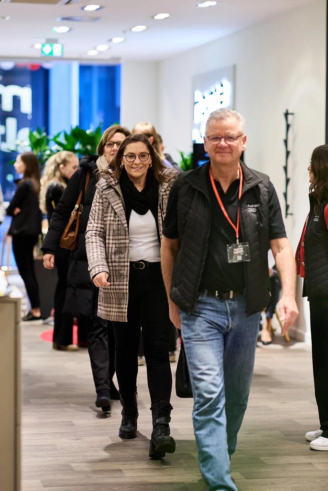Menschen gehen in einem modernen, hell erleuchteten Gebäude, vermutlich einer Messe oder einem Event, durch eine Garderobe. Im Vordergrund ist ein älterer Mann mit Brille und schwarzen Weste zu sehen, hinter ihm eine lächelnde Frau mit dunklen Haaren