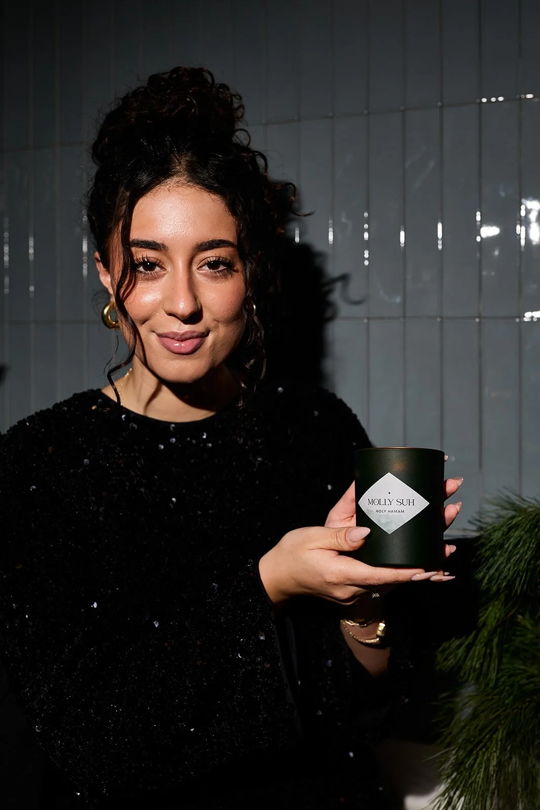 Eine Frau mit dunklen, lockigen Haaren in einem schwarzen, funkelnden Kleid hält eine Tasse mit dem Aufdruck 'MOLLY SUH HOLY HAMAM' in der Hand, im Hintergrund eine graue Wand mit vertikalen Fliesen und grüne Zweige.