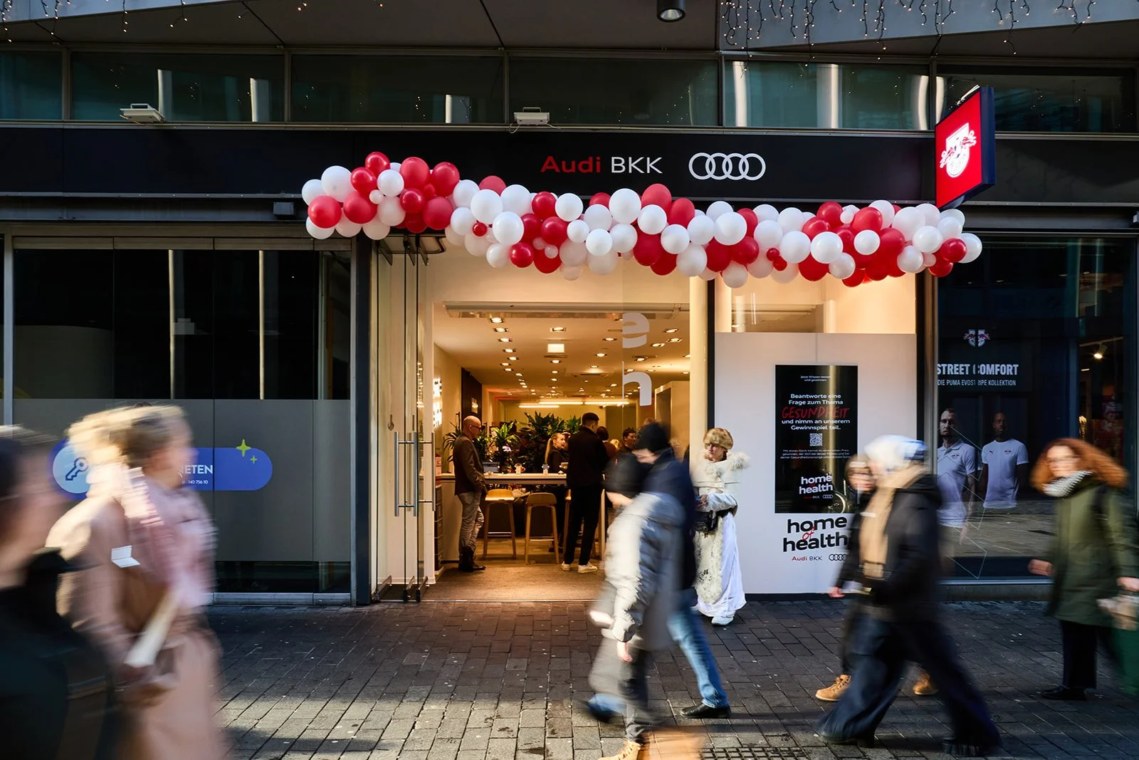 Eingang eines Geschäfts mit roten und weißen Ballons, Menschen gehen vorbei, im Inneren sieht man mehrere Personen an einem Empfangstresen, auf der rechten Seite eine digitale Anzeigetafel, Brandschilder von Audi, BKK und Home Health.