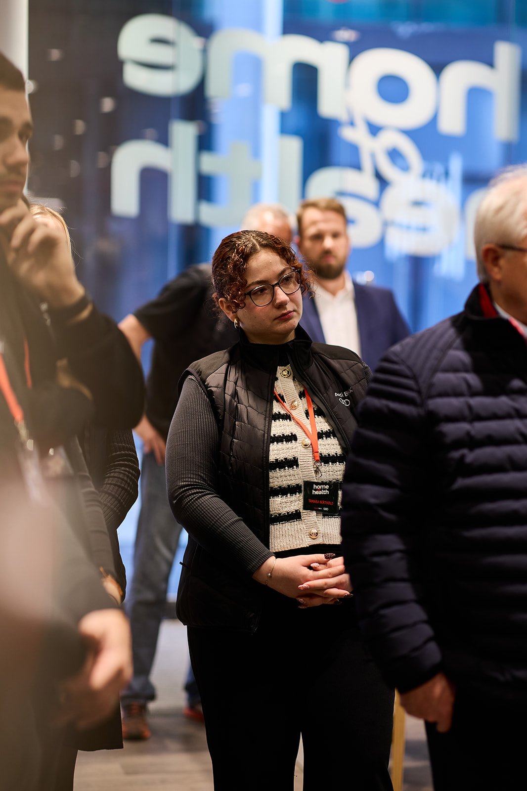 Menschen bei einer Konferenz oder Veranstaltung, eine Frau mit Brille trägt einen schwarzen Weste und einen namensschild, im Hintergrund blauer Hintergrund mit weißen, großen, unscharfen Buchstaben.
