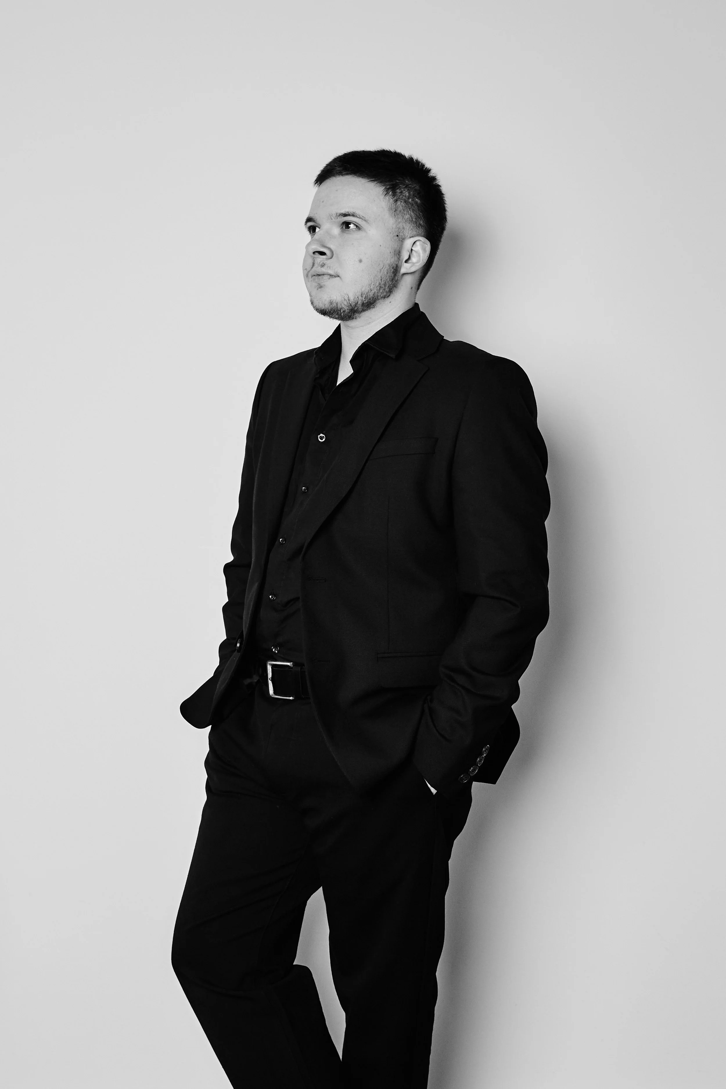 Mann im schwarzen Anzug lehnt an weißer Wand, Blick nach links, Hand in Tasche, Schwarz-Weiß-Foto.