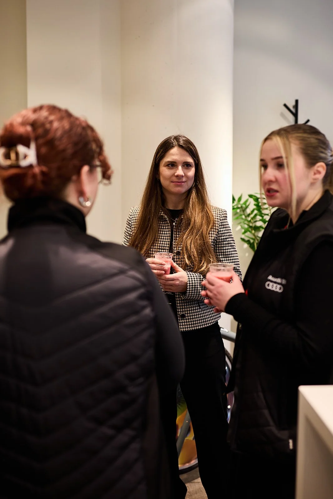 Drei Frauen stehen im Gespräch und trinken Getränke in einem Raum mit Pflanzen, weißer Wand und neutraler Beleuchtung.