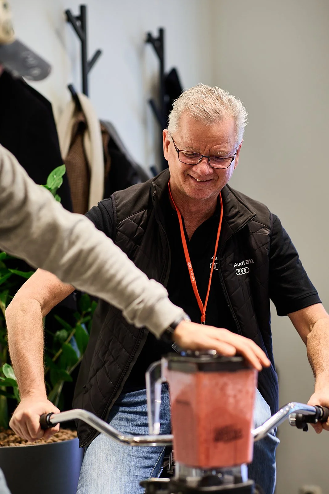 Ein älterer Mann mit Brille und grauen Haaren, der auf einem Fahrrad sitzt, während jemand anderes ihm den Halter am Lenker hält. Der Mann trägt ein schwarzes T-Shirt mit einem Audi-Logo und einer schwarzen Weste.