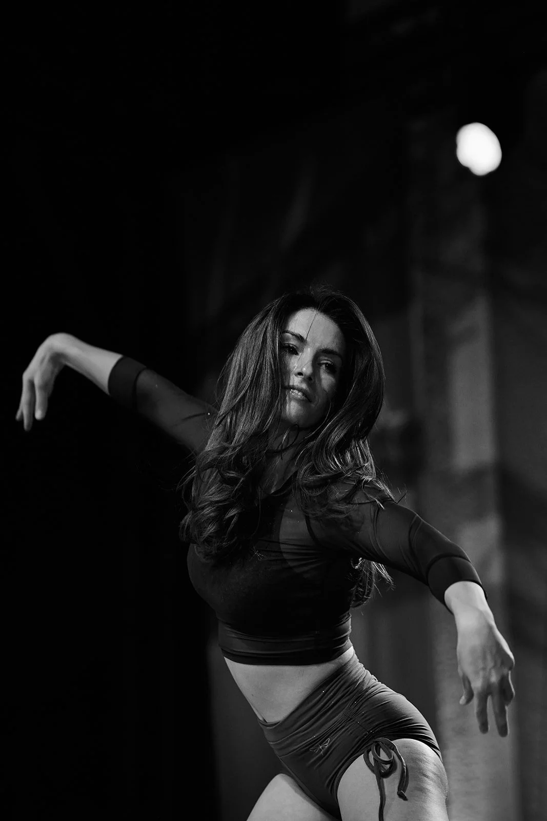 Eine junge Frau tanzt in einem dunklen Raum, trägt ein schwarzes Sportoberteil und Shorts, mit langem, lockigem Haar.