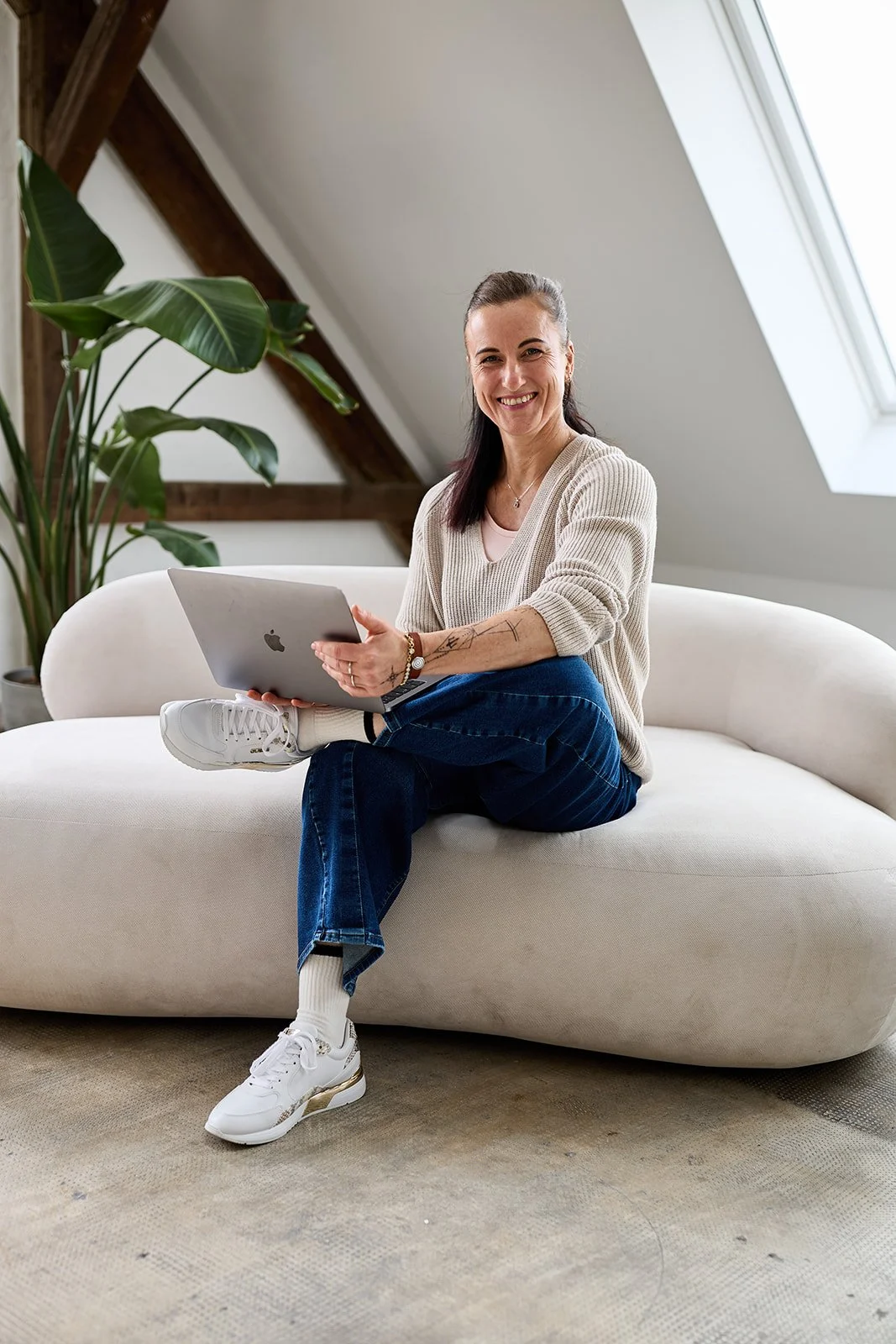 Eine lächelnde Frau sitzt auf einer weißen Couch im Dachgeschoss, hält einen Laptop in den Händen, trägt einen cremefarbenen Pullover, blaue Jeans und weiße Sneakers, im Hintergrund eine große Pflanze und ein Fenster an der schrägen Zimmerdecke.