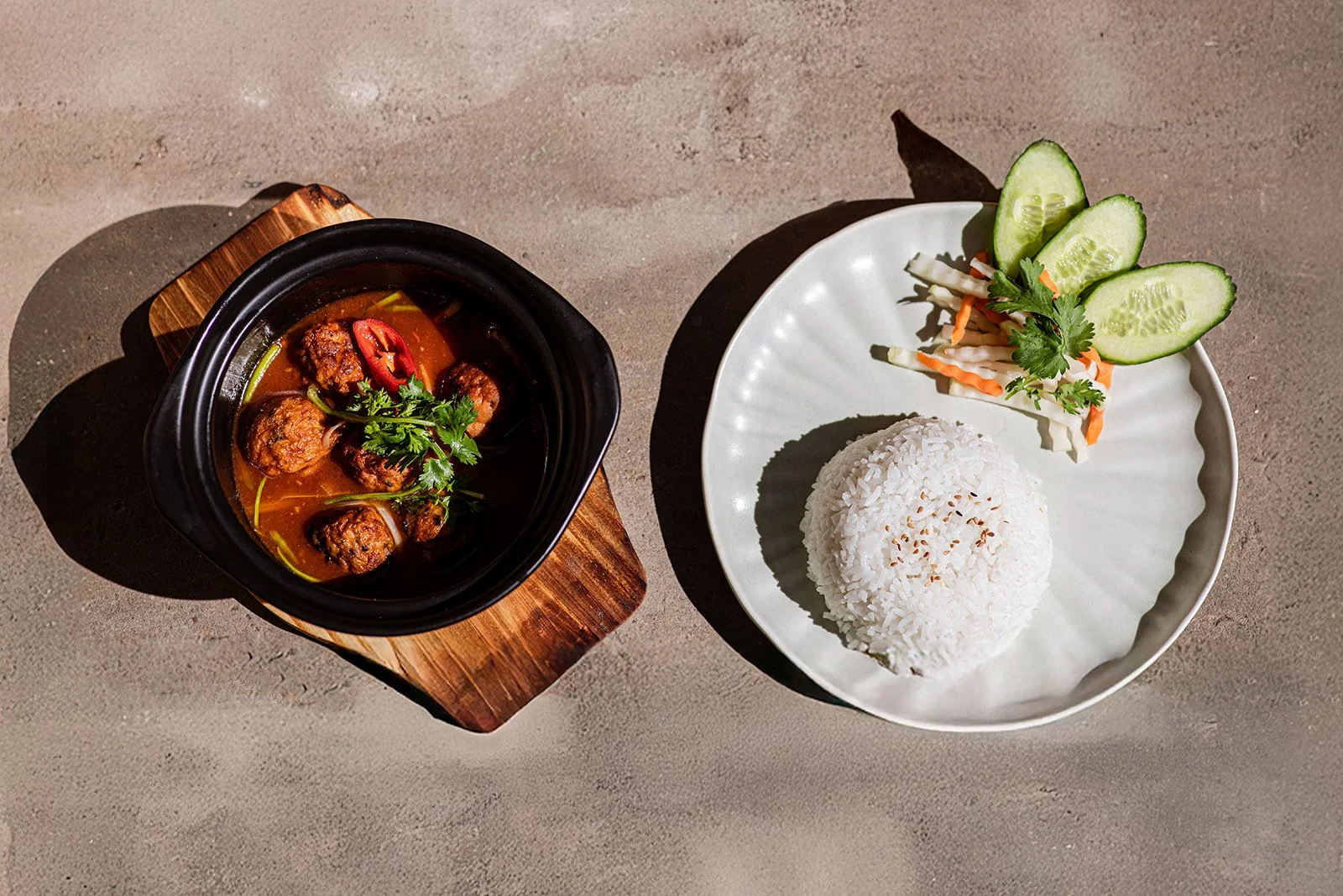 Ein Teller mit Reis, Gurkenscheiben, Karotten und Koriander sowie ein Topf mit Fleischbällchen in einer braunen Soße