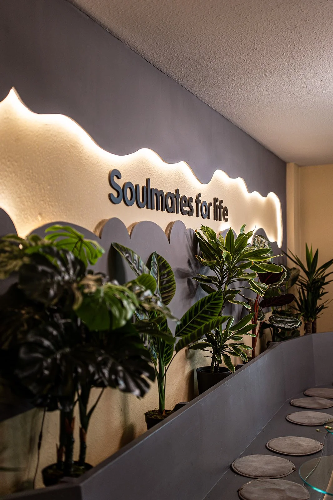 Innenansicht eines Raumes mit einem Wandrelief, das den Text "Soulmates for life" zeigt, umgeben von Pflanzen und einem Esstisch mit runden Platzdeckchen.