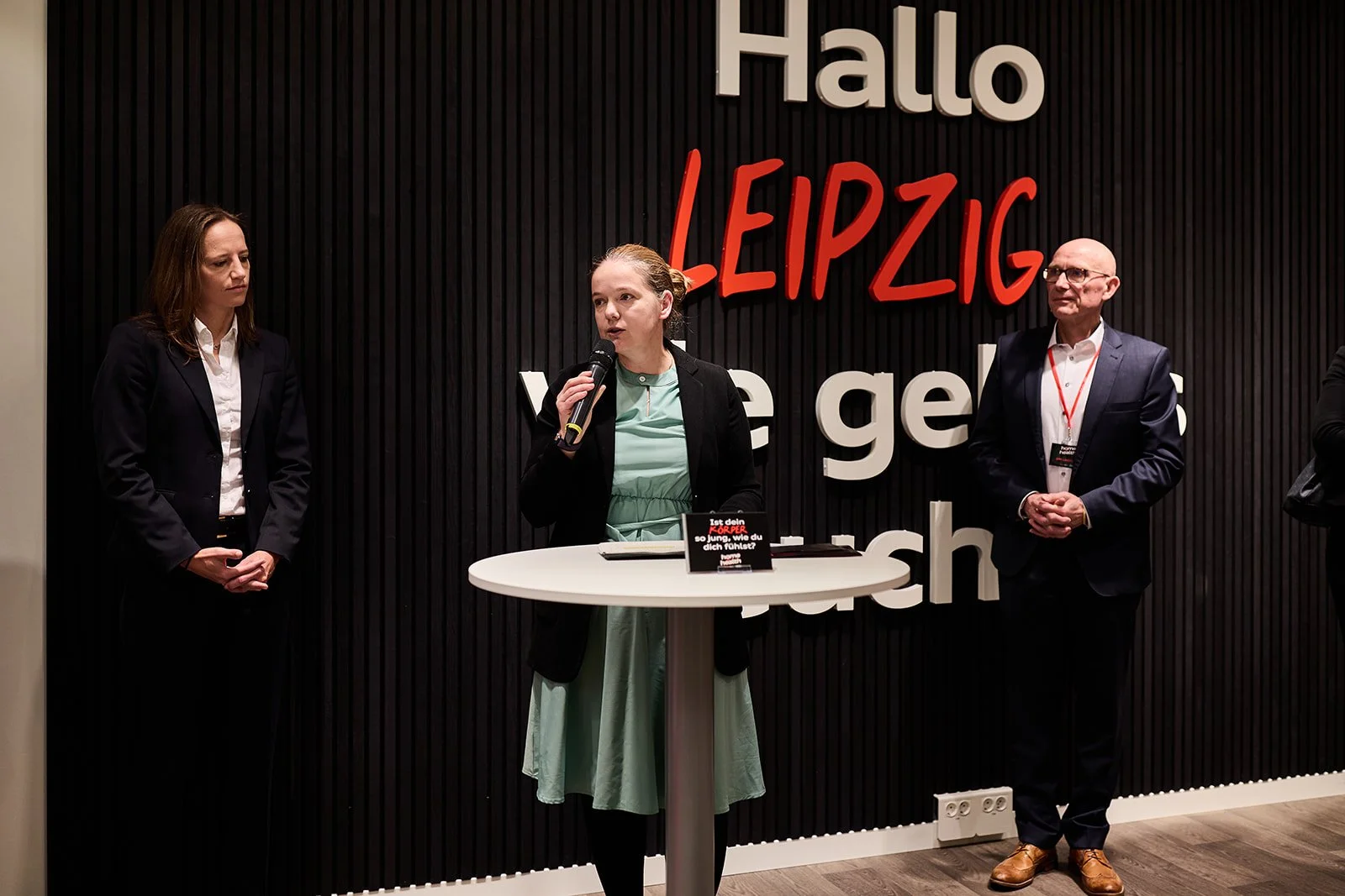 Drei Personen stehen vor einer schwarzen Wand mit großen weißen und roten Buchstaben, die 'Hallo Leipzig wie geht's' sagen. Eine Frau in der Mitte spricht mit einem Mikrofon, während die anderen beiden zuhören.