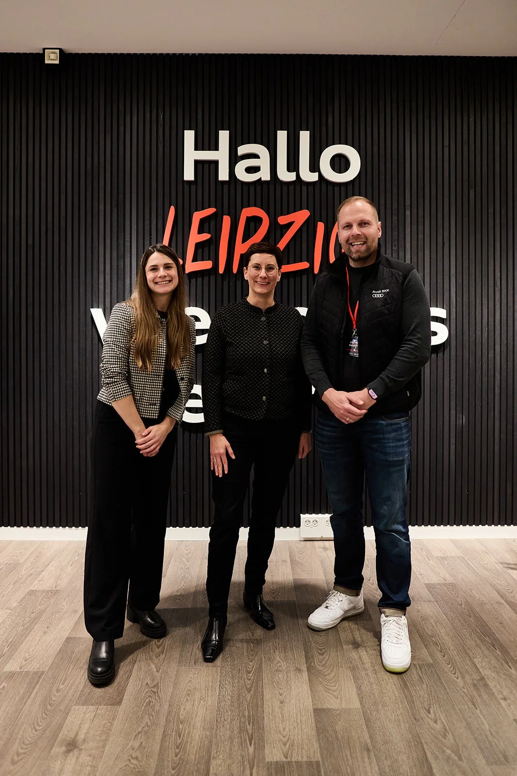 Drei Personen stehen vor einer schwarzen Wand mit der Aufschrift "Hallo LEIPZIG" in weißen und roten Buchstaben. Die Person auf der linken Seite ist eine Frau mit langen braunen Haaren, die eine karierte Jacke trägt, die mittlere Person ist eine Frau
