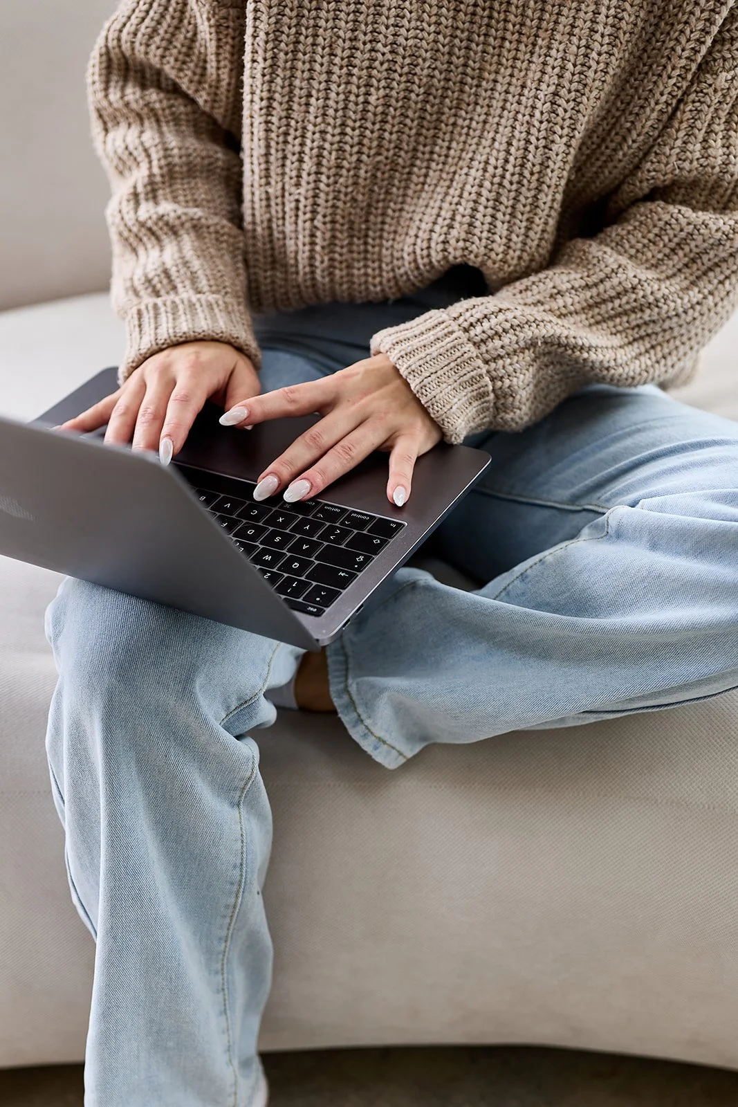Person sitzt auf einem Sofa mit einem Laptop auf den Knien, trägt einen beigen Strickpulli und blue Jeans.