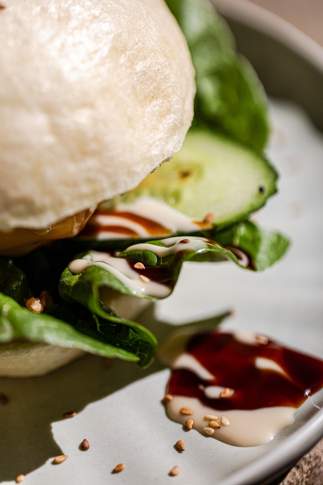 Nahaufnahme eines Salats mit Gurke, Eisbergsalat, zwei Dressings (weißer und brauner), Sesamsamen und einer Scheibe Kohlsalat auf einem weißen Teller.