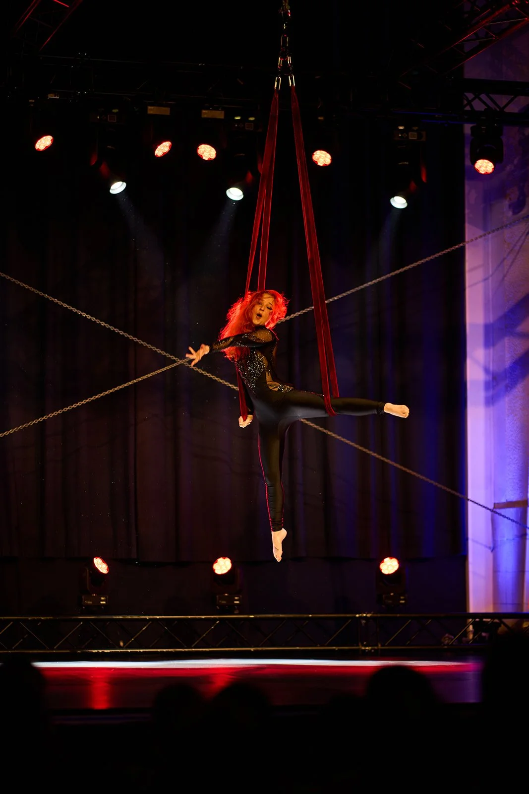 Poletänzerin auf einem Aerial Hoop im Theater, bei rotem Bühnenlicht, schwarzem Bühnenhintergrund und dramatischer Beleuchtung.