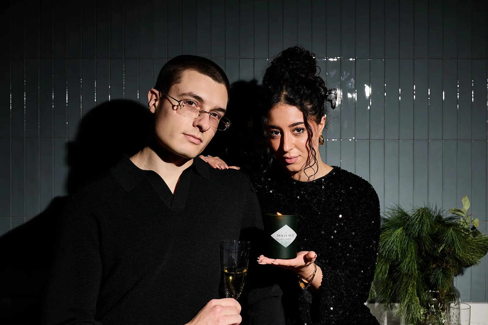 Ein Mann und eine Frau feiern zusammen, der Mann hält ein Glas Champagner, die Frau hält eine Kerze mit dem Label 'Molly Sue', sie tragen schwarze Kleidung, im Hintergrund sind grüne Pflanzen und eine dunkle Wand.