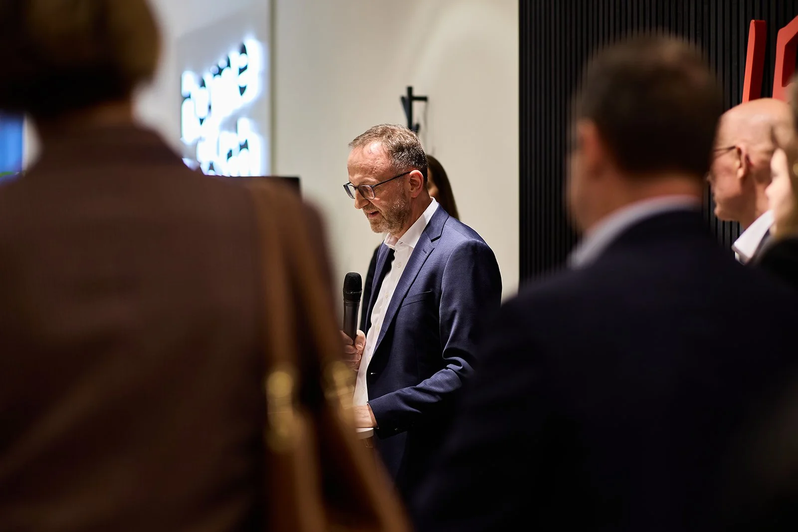 Ein Mann in einem blauen Anzug mit Brille hält ein Mikrofon in der Hand und spricht vor einer Gruppe von Menschen bei einer Veranstaltung.