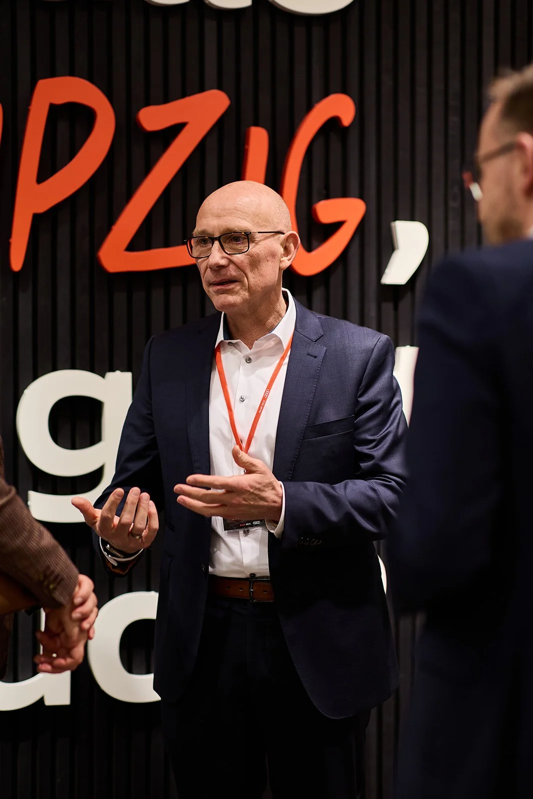 Ein älterer Mann in Anzug mit Brille spricht auf einer Veranstaltung, hinter ihm ist ein schwarzer Hintergrund mit roten und weißen Buchstaben, die teilweise den Begriff 'PZICK' bilden.